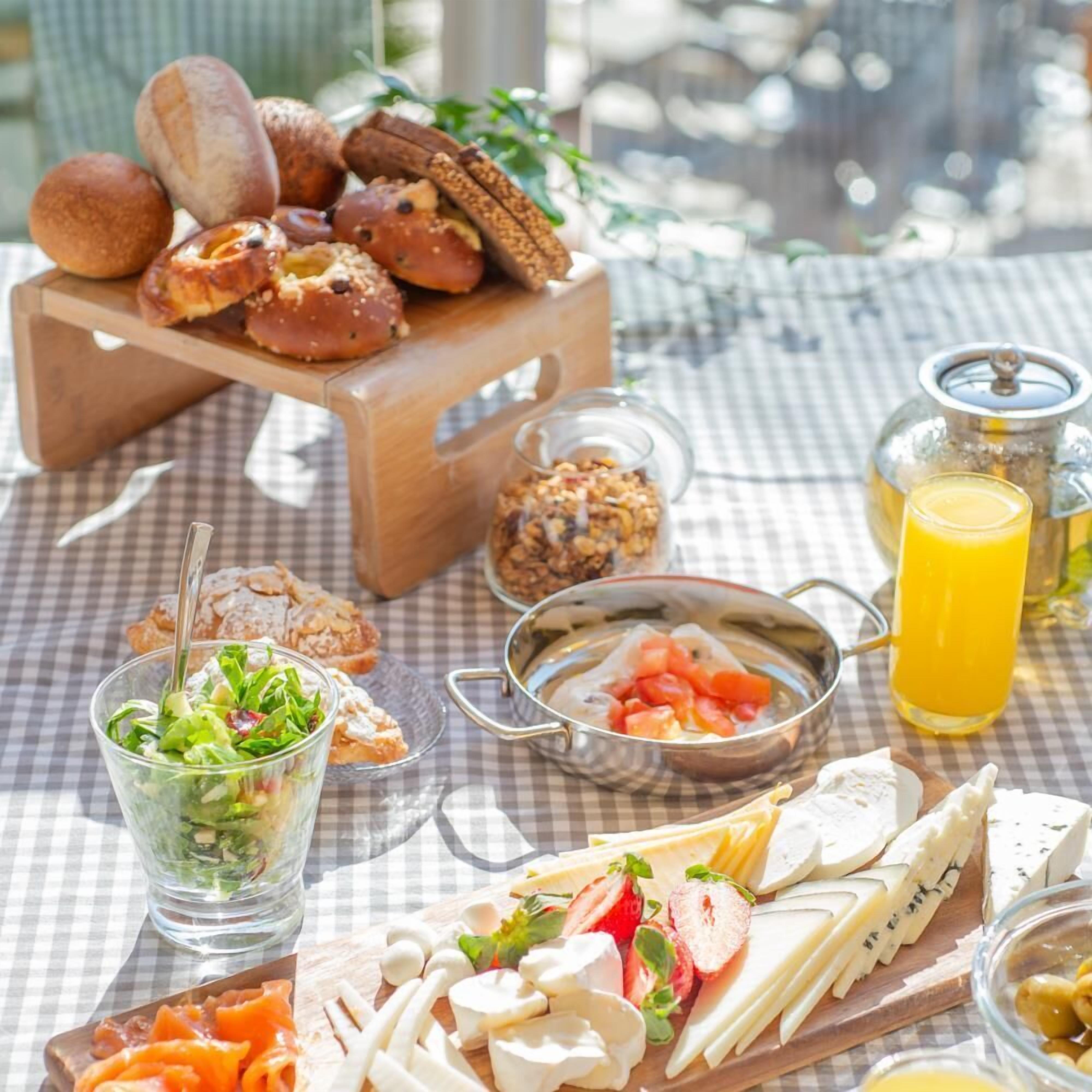 Colazione servita con pane, pasticcini, formaggio, frutta e un bicchiere di succo d'arancia su una tovaglia a quadri.