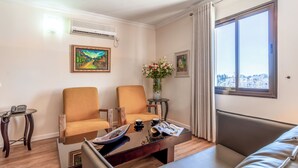 Une chambre d’hôtel moderne, dotée de deux fauteuils, d’un canapé, d’une table basse et offrant une vue sur la ville grâce à une grande fenêtre.