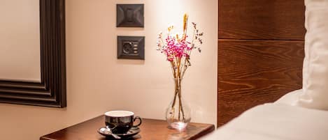 A neatly arranged hotel room with a wooden nightstand, a bedside table with a cup and saucer, a vase with flowers, and a bed with white bedding.
