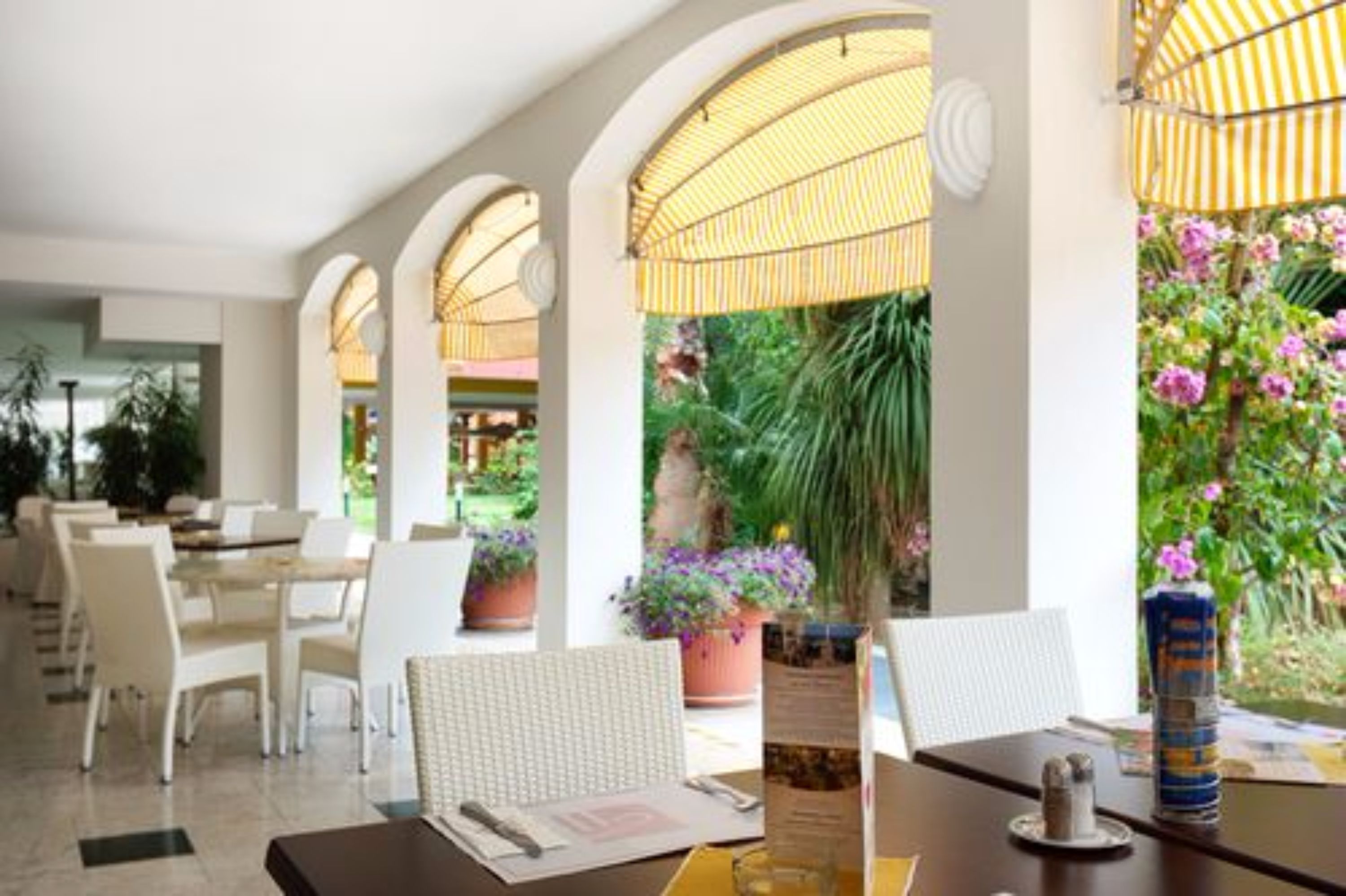 Un restaurant avec des chaises et des tables blanches, un présentoir à menus et une vue sur un jardin agrémenté de plantes et de fleurs.
