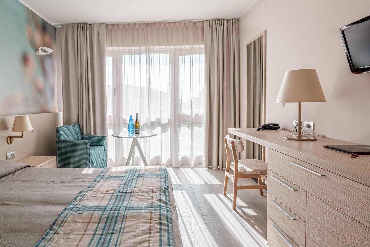 Quarto de hotel com cama, escrivaninha, cadeira, televisão e janela com cortinas.