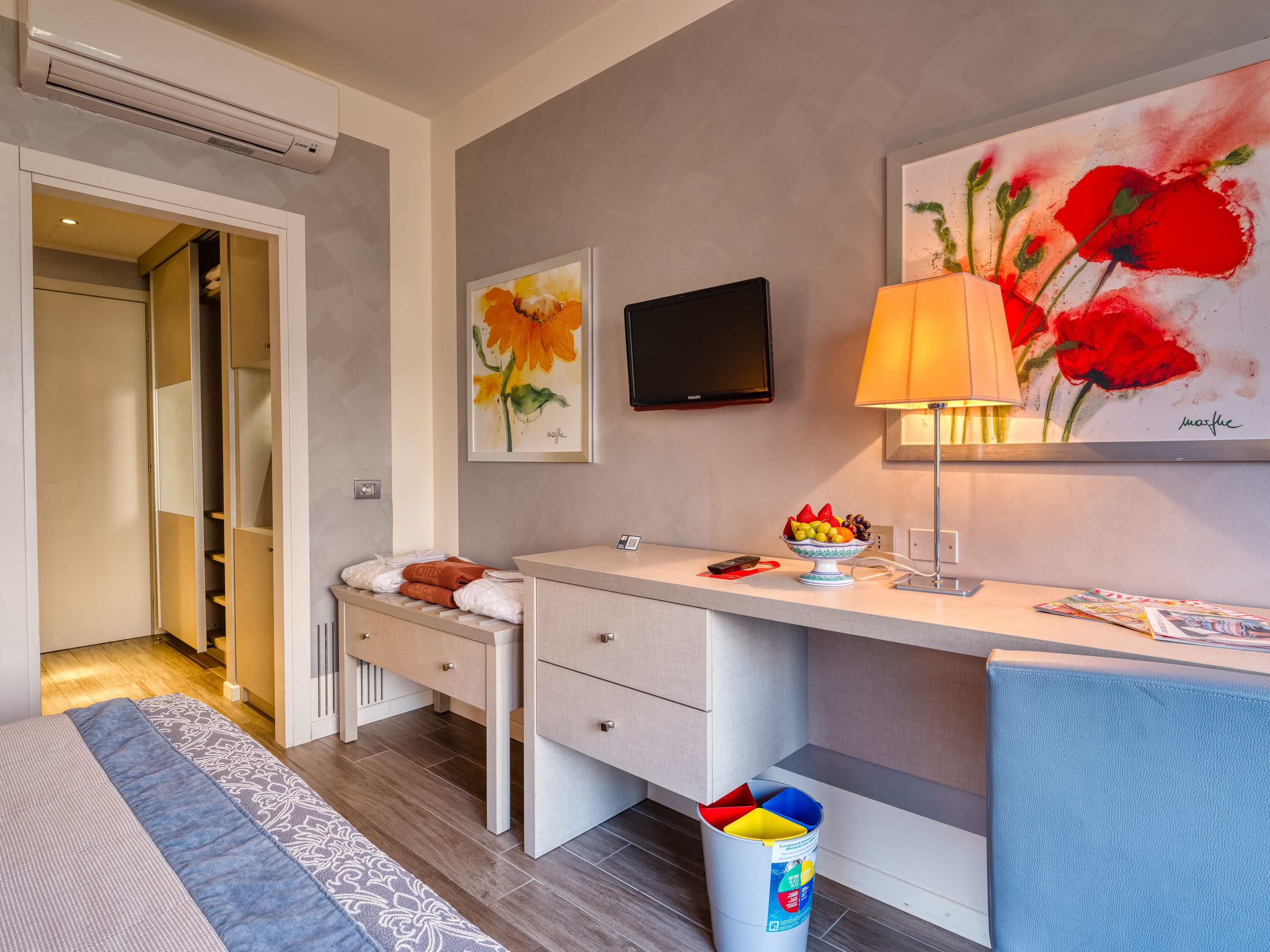 A modern hotel room with a bed, a desk with a lamp, a TV, and a painting of flowers.