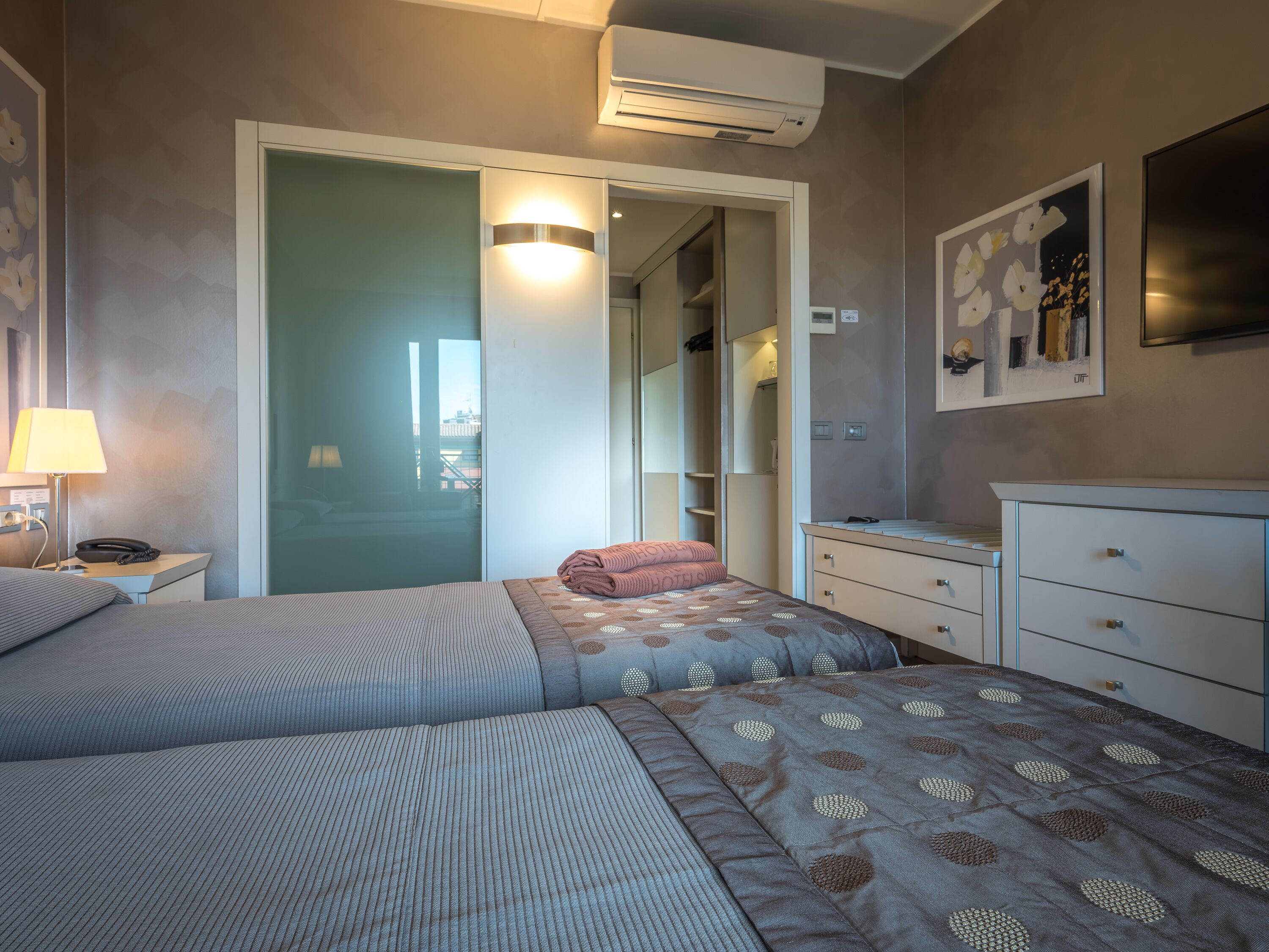 Ein Hotelzimmer mit zwei Betten, einem Schreibtisch, einer Lampe, einem Kleiderschrank und einem Bild an der Wand.