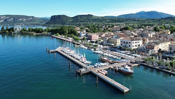 Un puerto deportivo con barcos amarrados, una noria y un pueblo con edificios y zonas verdes.