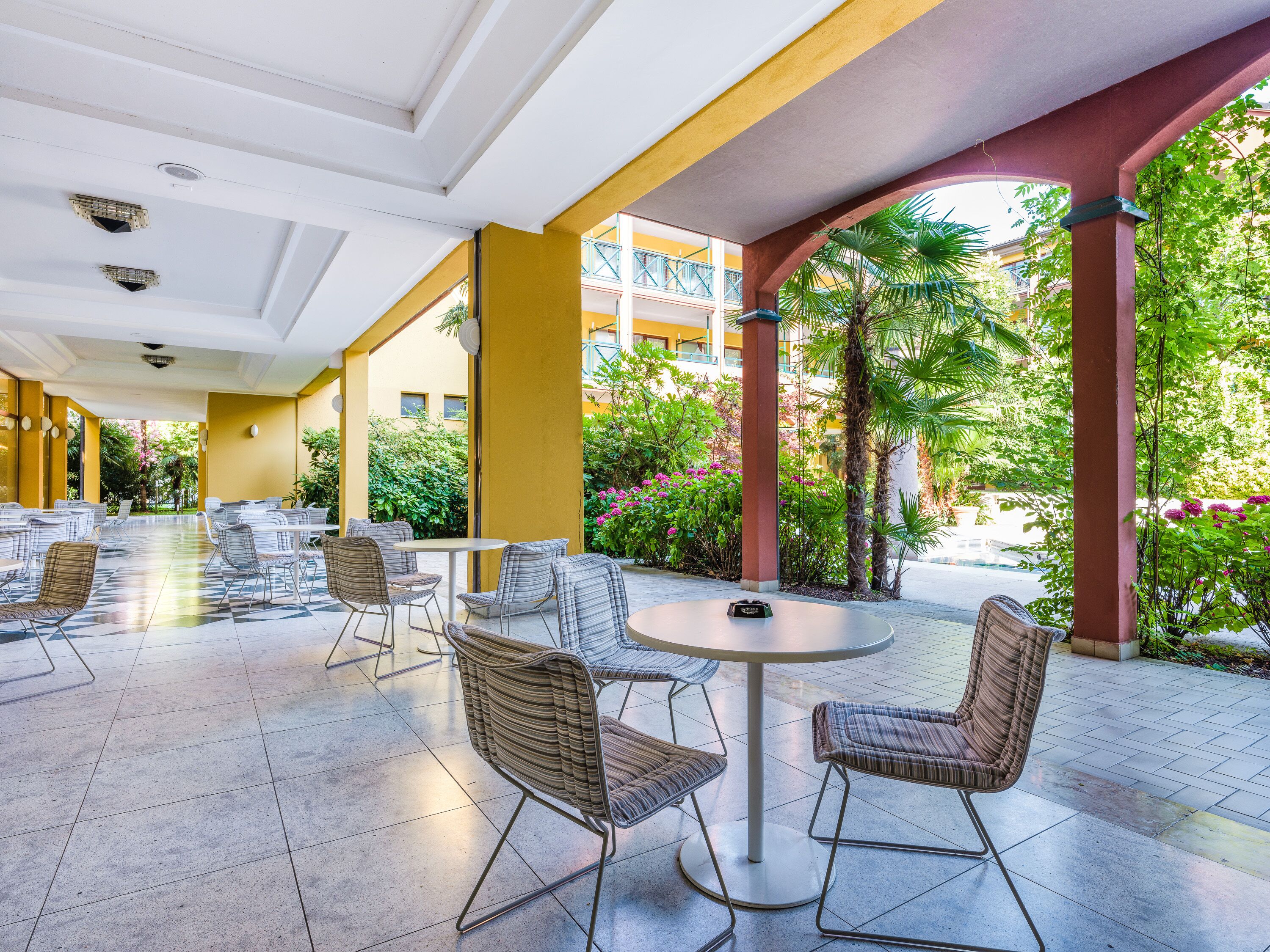 Une vaste terrasse extérieure dotée de plusieurs tables et de chaises en osier, entourée de verdure et offrant une vue sur un bâtiment en arrière-plan.