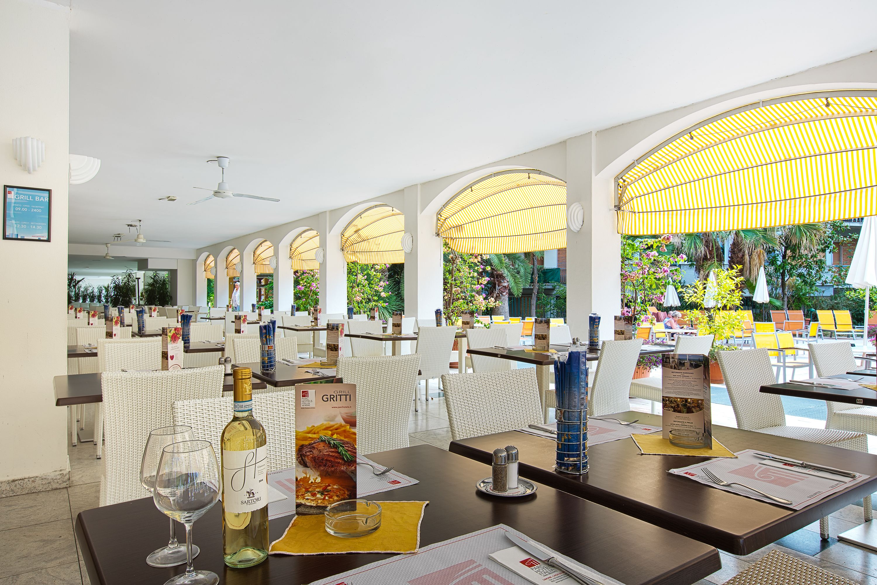 Ein Restaurant mit weißen Korbstühlen, Tischen mit Speisekarten und Blick ins Freie.