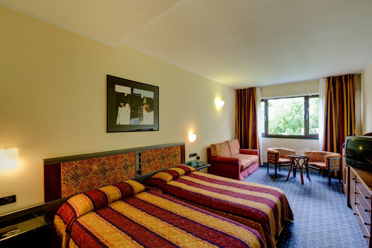 Quarto de hotel com duas camas, um sofá, uma televisão e uma janela com cortinas.