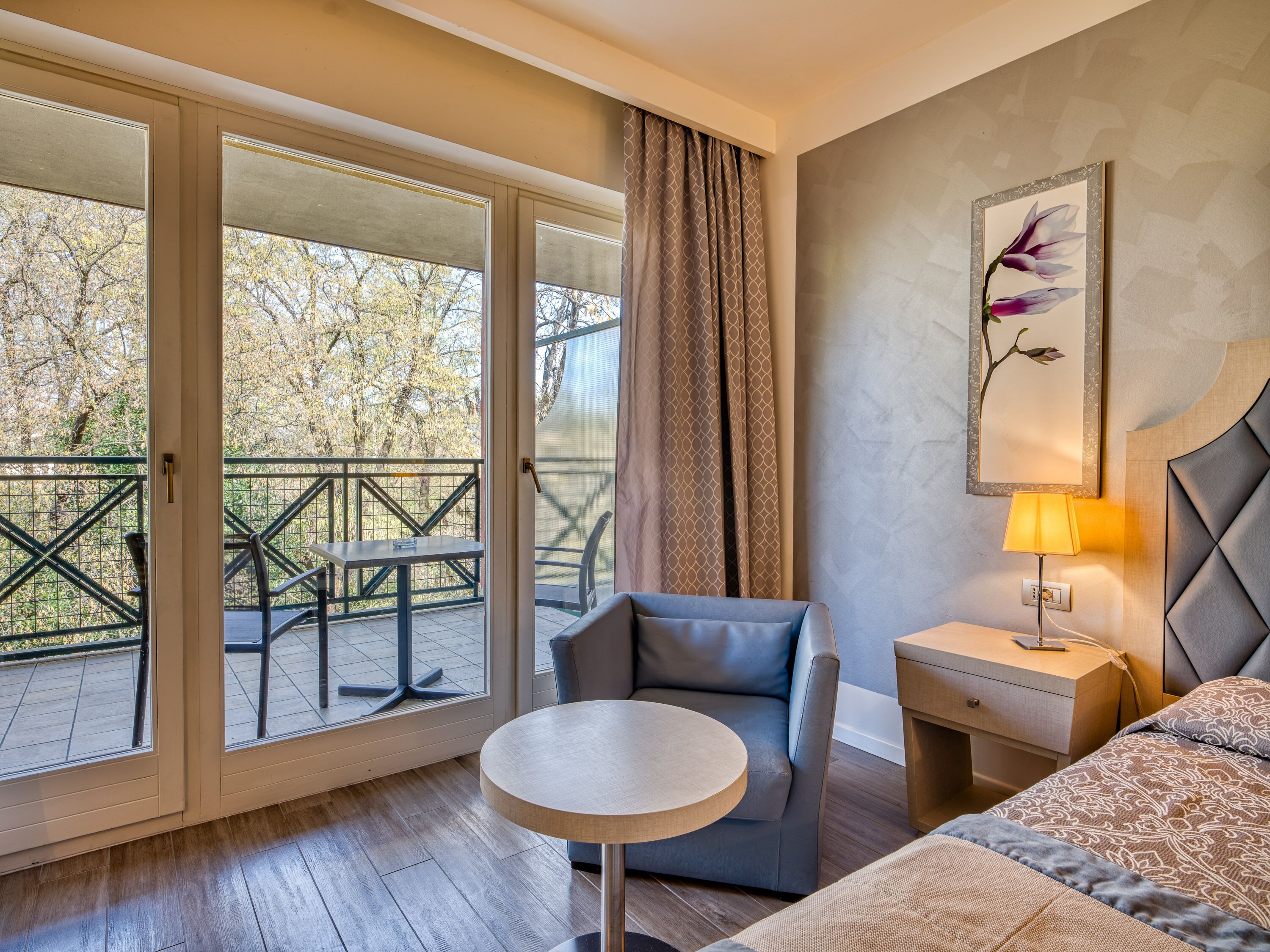 A hotel room with a bed, a round table, a chair, a nightstand, and a balcony with a table and chairs.