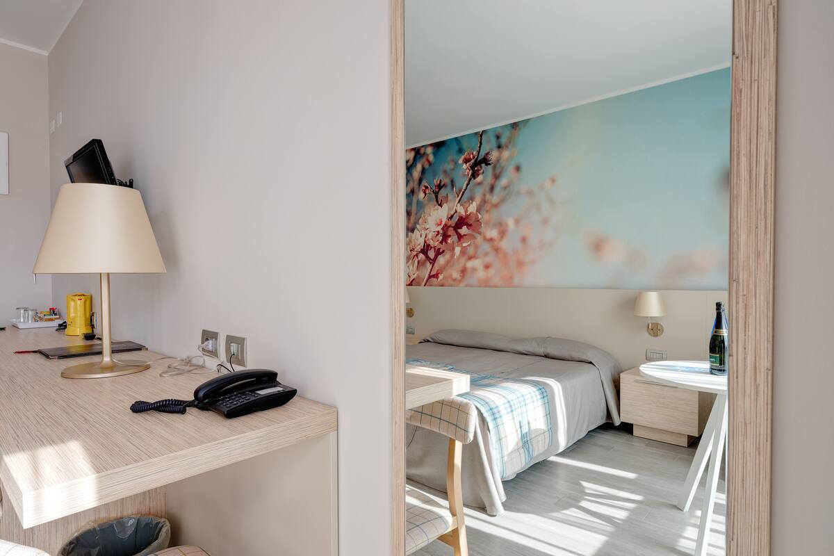 Quarto de hotel com escrivaninha, luminária, telefone, lixeira, cama com manta xadrez e parede com pintura floral.