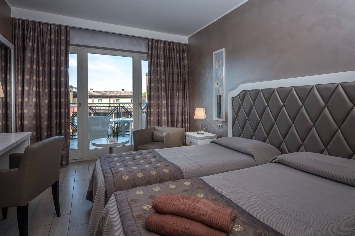 Quarto de hotel com cama, escrivaninha e cadeira. Há uma janela com cortinas e vista para edifícios.