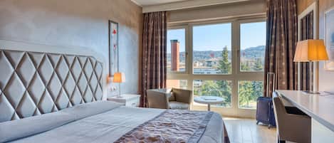 A hotel room with a large bed, a desk, a chair, and a view of the city through large windows.