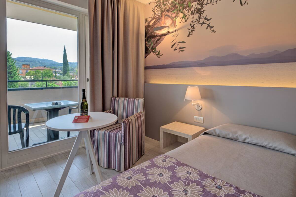 Quarto de hotel com cama, uma mesinha com uma garrafa, uma cadeira, um criado-mudo e vista para o exterior.