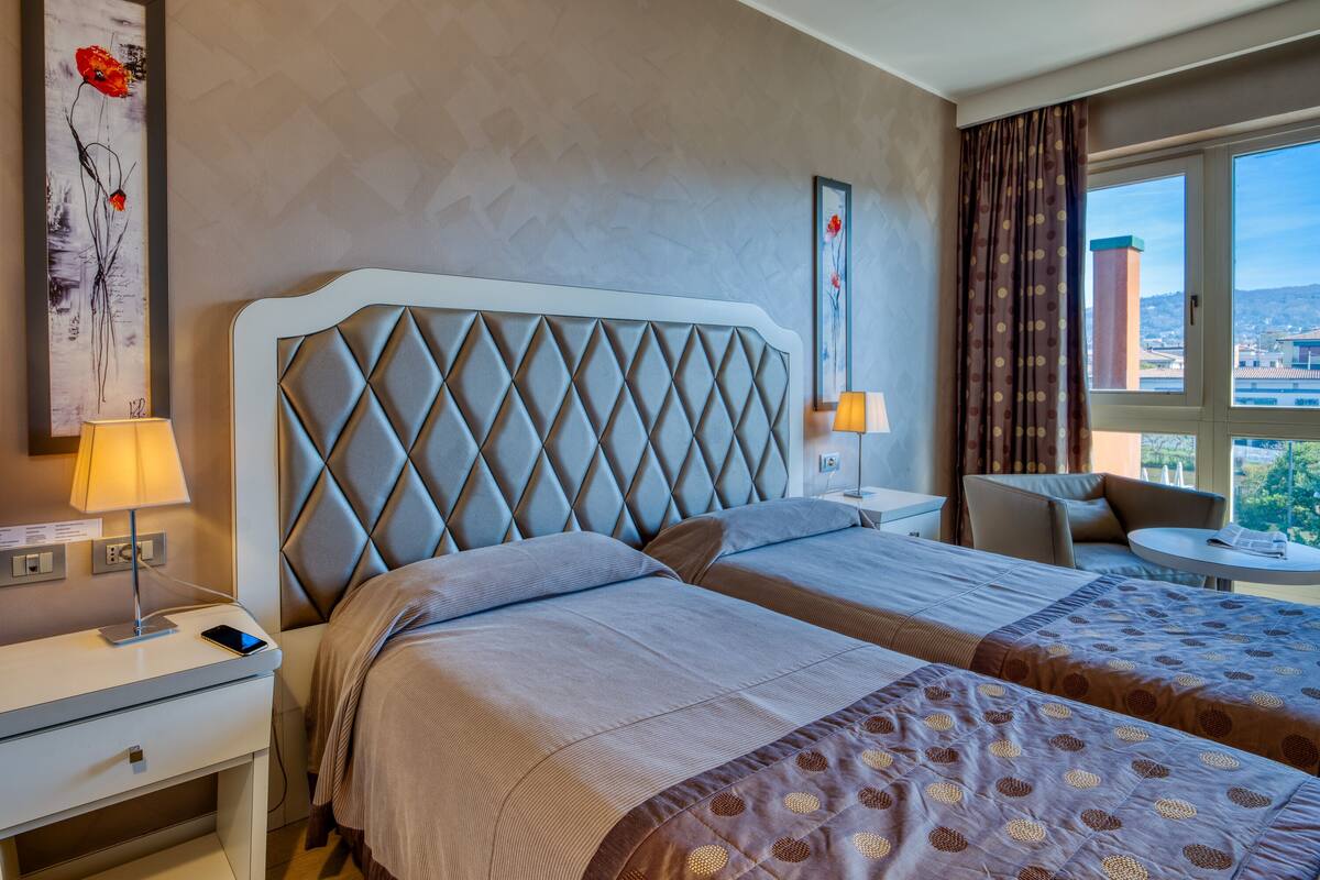 Quarto de hotel com uma cama grande, duas mesinhas de cabeceira, uma cadeira e vista para a cidade.
