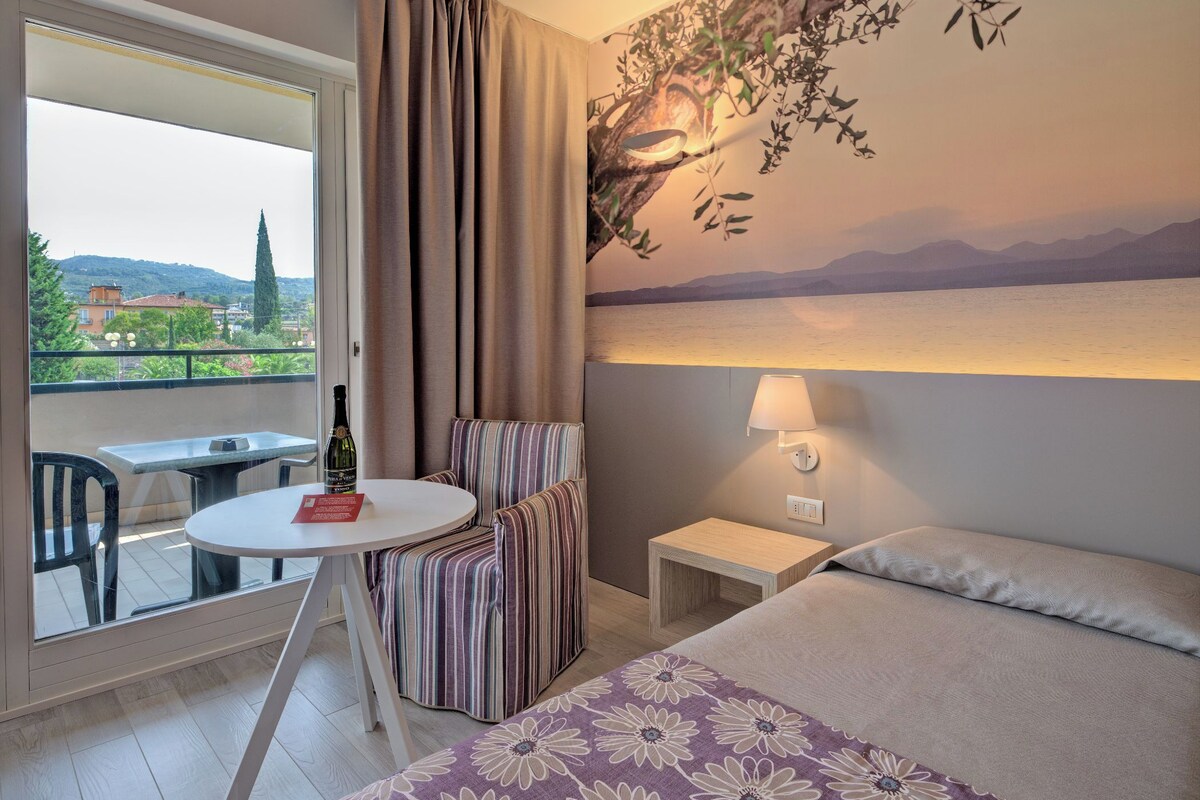 Una habitación de hotel con una cama, un sillón a rayas, una mesita con una botella, una mesita de noche y una lámpara de pared.