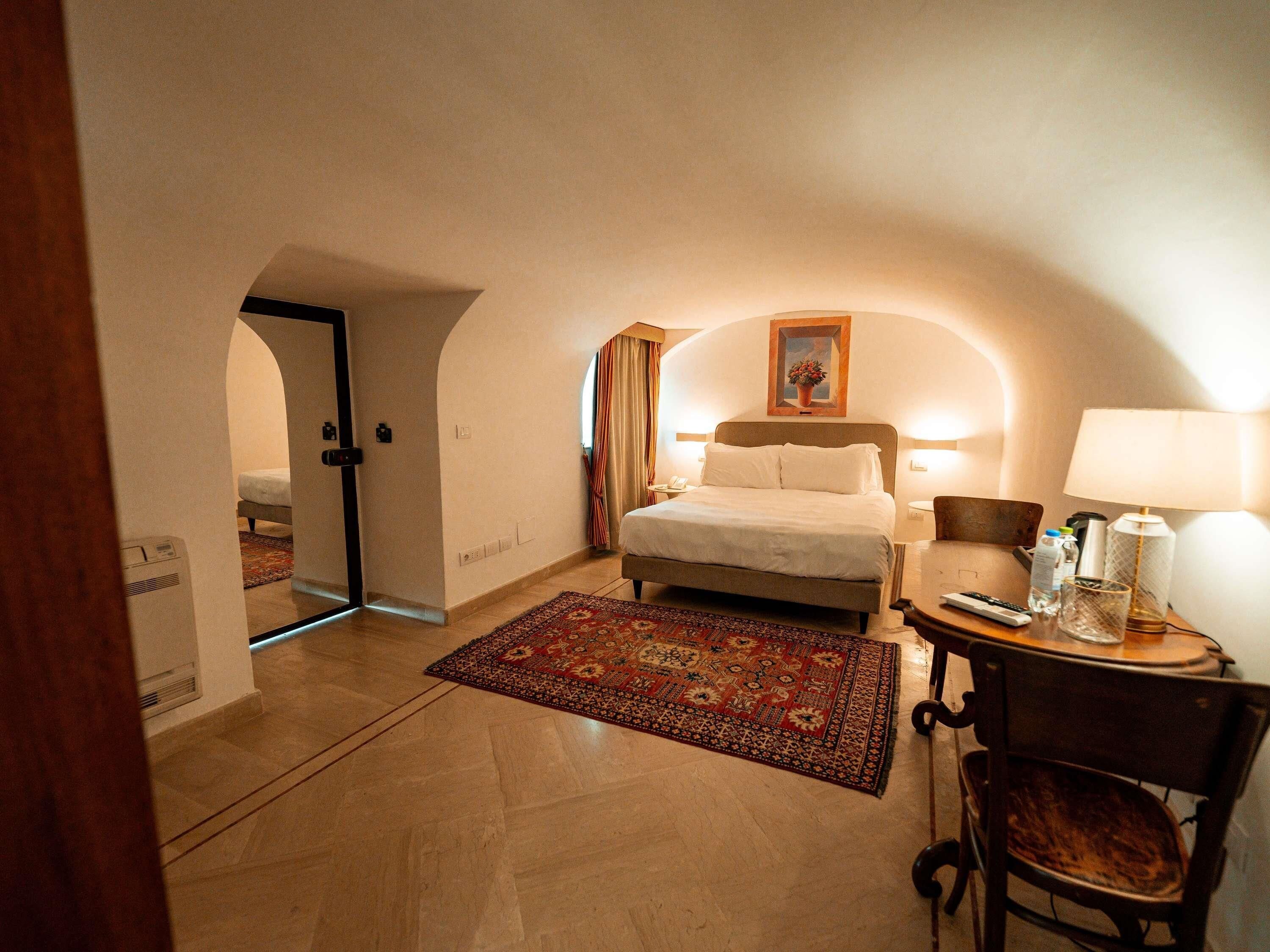 A hotel room with a bed, a desk with a lamp, a chair, a rug, and a painting on the wall.