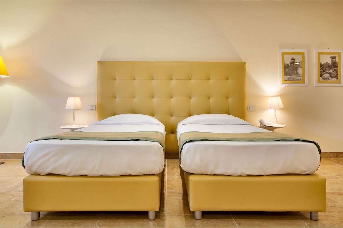A hotel room with two beds, a tufted headboard, bedside lamps, and two framed pictures on the wall.