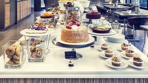 A buffet with various desserts and pastries, including cakes and muffins, in a modern dining area with a kitchen in the background. - Mercure Villa Romanazzi Carducci Bari (Bari)