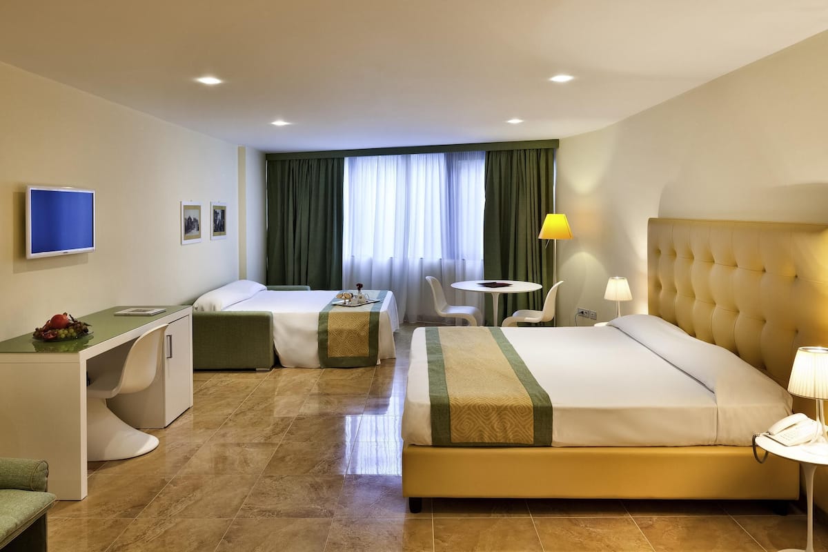A hotel room with a bed, a desk, a chair, a small table, a TV, and a window with curtains.