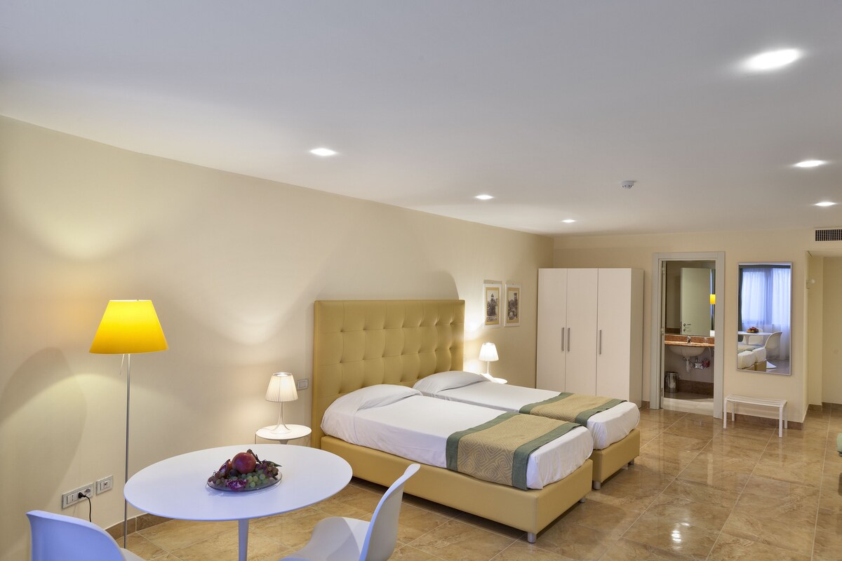A hotel room with two beds, a round dining table, a small white bench, and a bathroom visible through an open door.