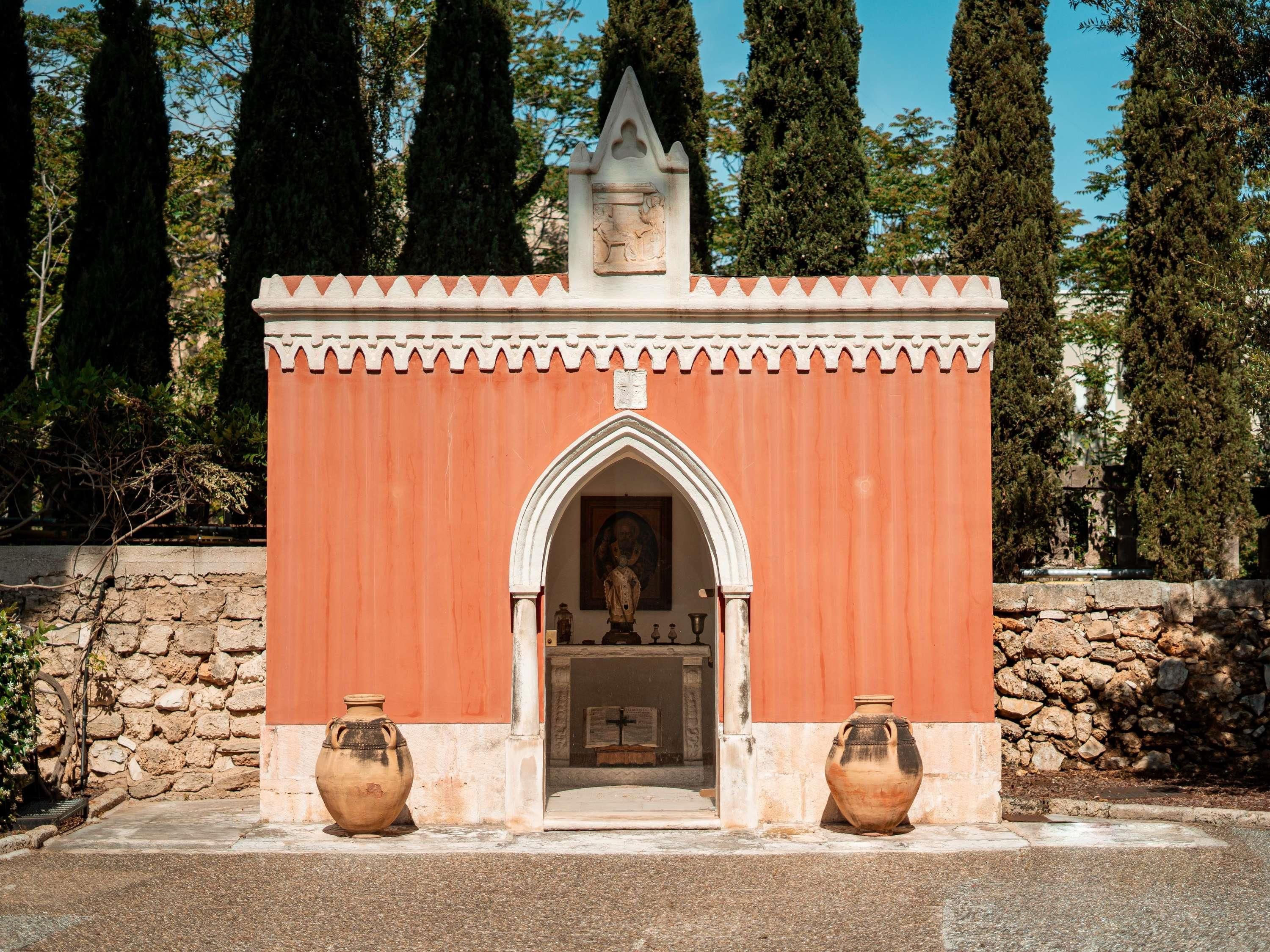 A small chapel with a red facade, a pointed arch entrance, and two large vases at the front.