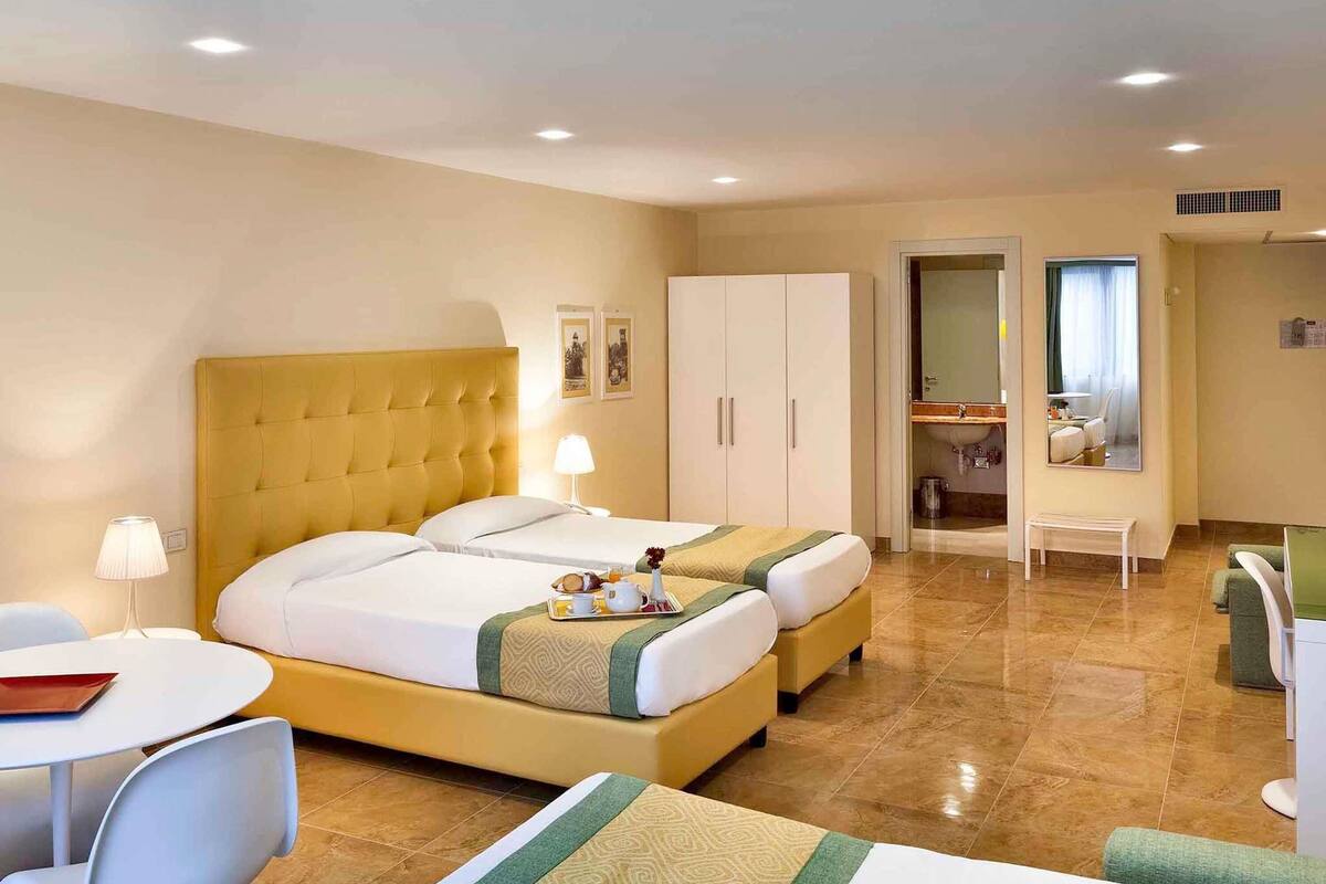 Camera d'albergo con due letti, una scrivania, una sedia, uno specchio e un bagno visibile attraverso una porta aperta.