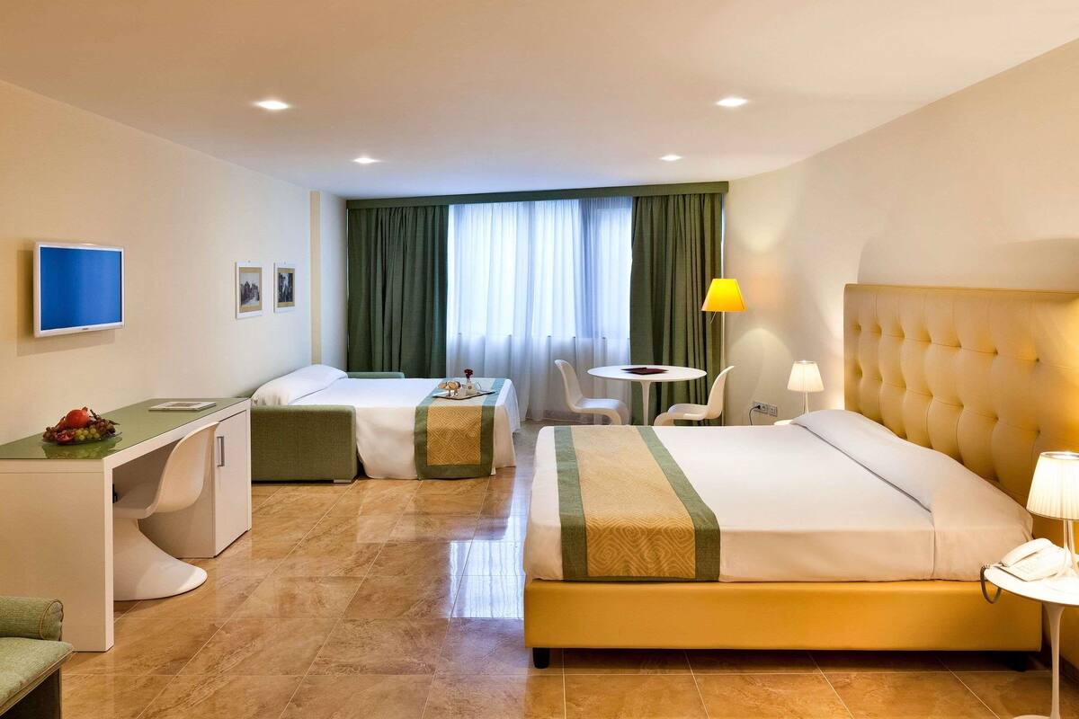 Camera d'albergo con un letto, una scrivania, una sedia, un tavolino, una televisione e una finestra con tende.