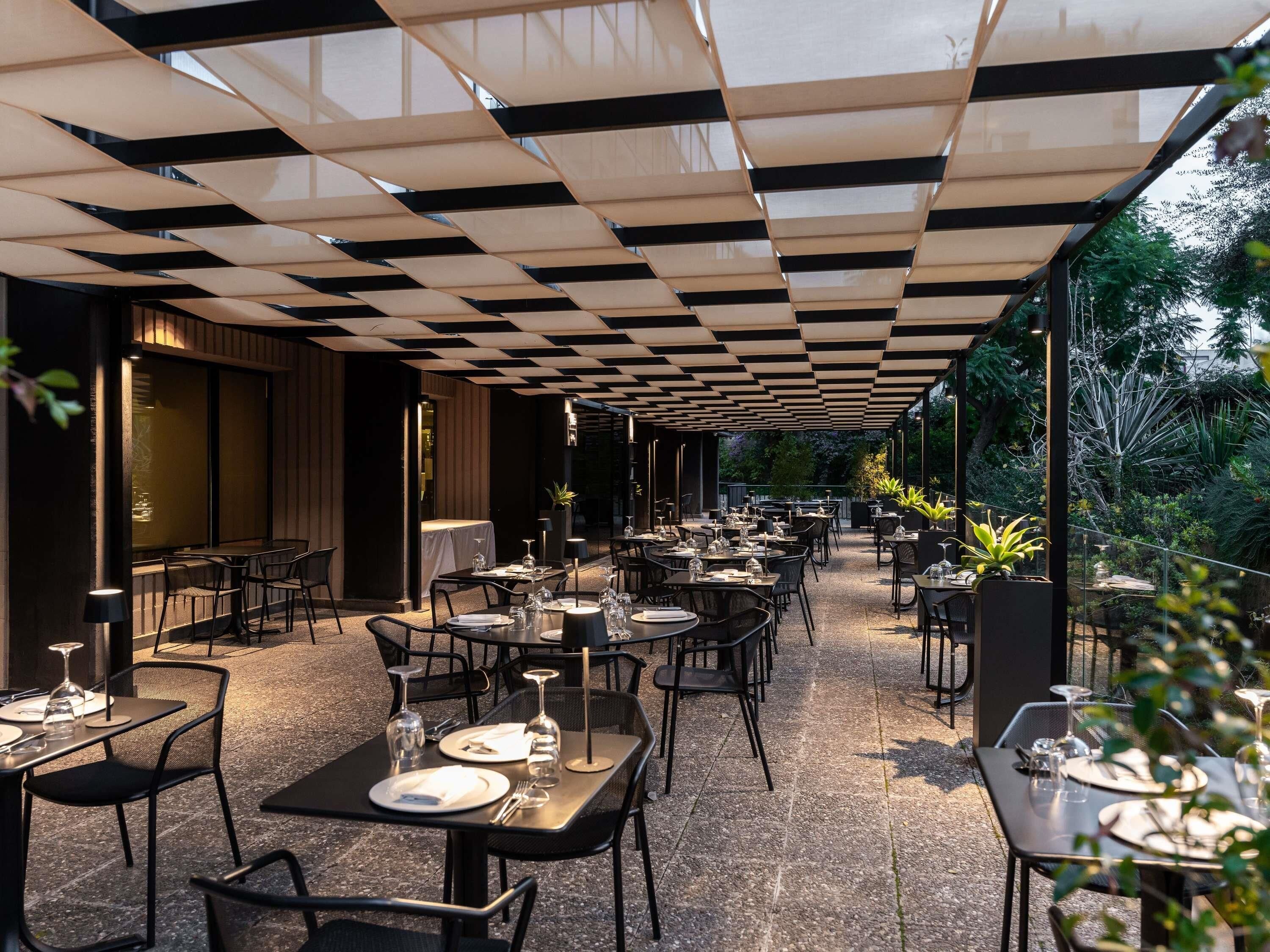 Un área de comedor al aire libre con mesas y sillas, techo a cuadros y plantas en macetas.