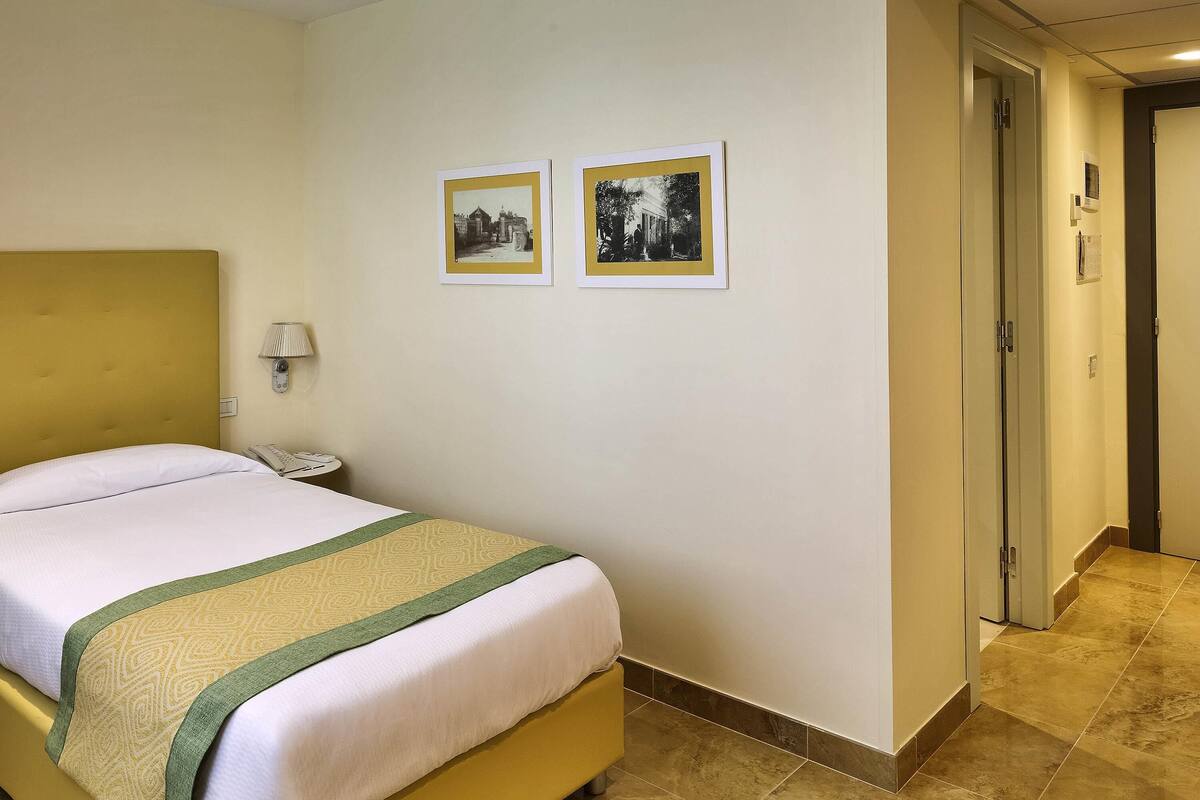 Una camera d'albergo con un letto, un telefono sul comodino, una lampada e due quadri appesi al muro.