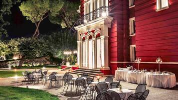 An outdoor dining area with tables and chairs, a red building with a balcony, and trees in the background.