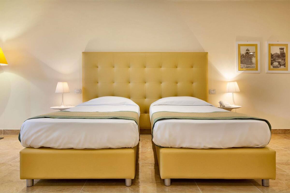 A hotel room with two beds, a tufted headboard, bedside lamps, and two framed pictures on the wall.