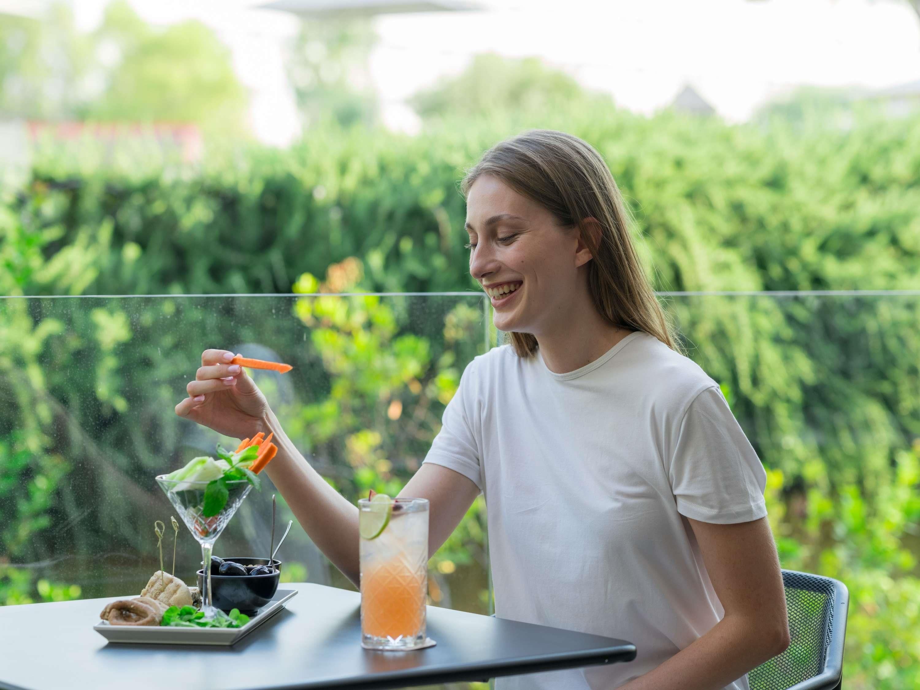 Una persona sentada a una mesa con una bebida y comida, al aire libre con follaje de fondo.