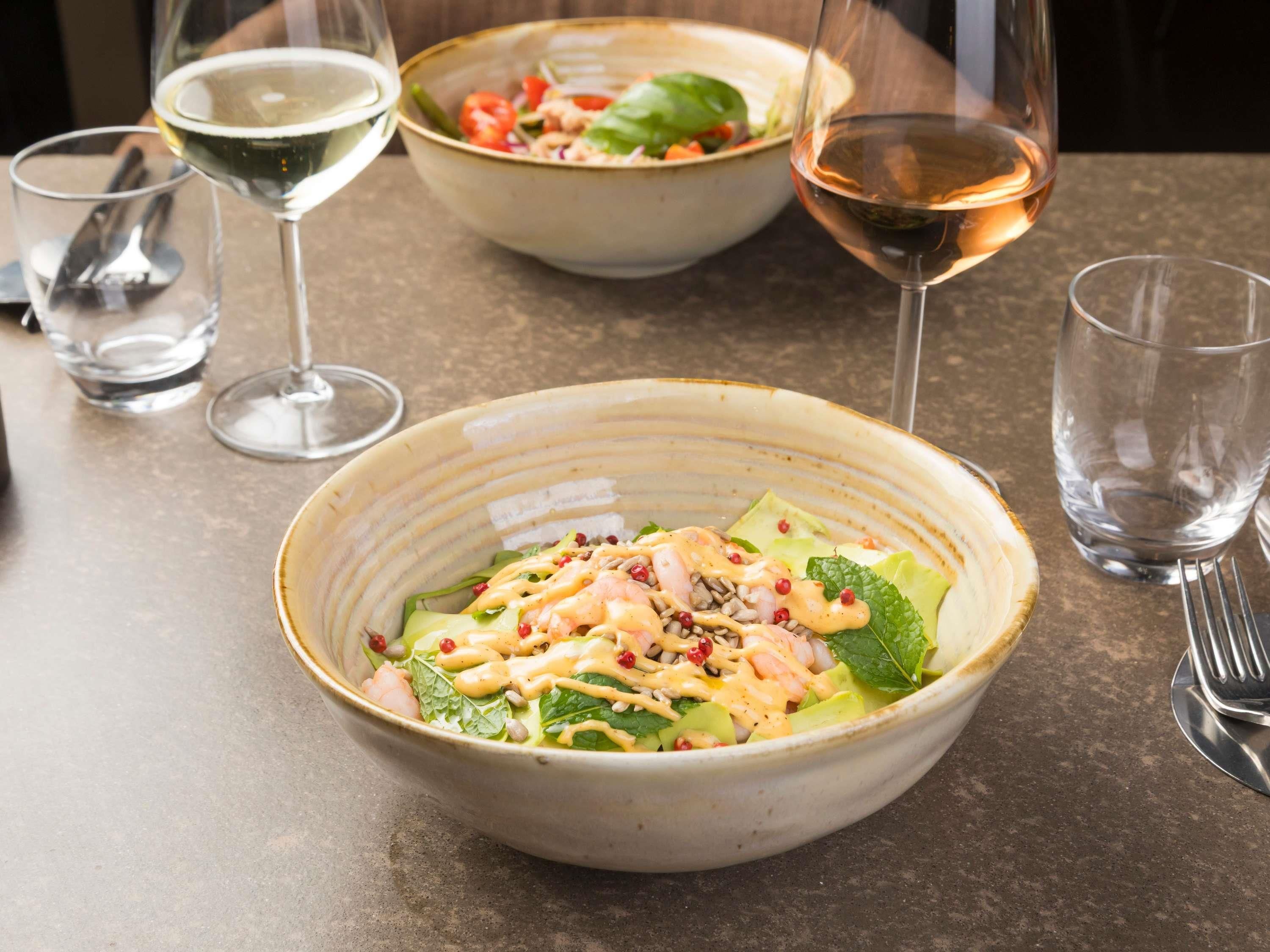 A dining table with a bowl of salad, two glasses of white wine, and a fork.