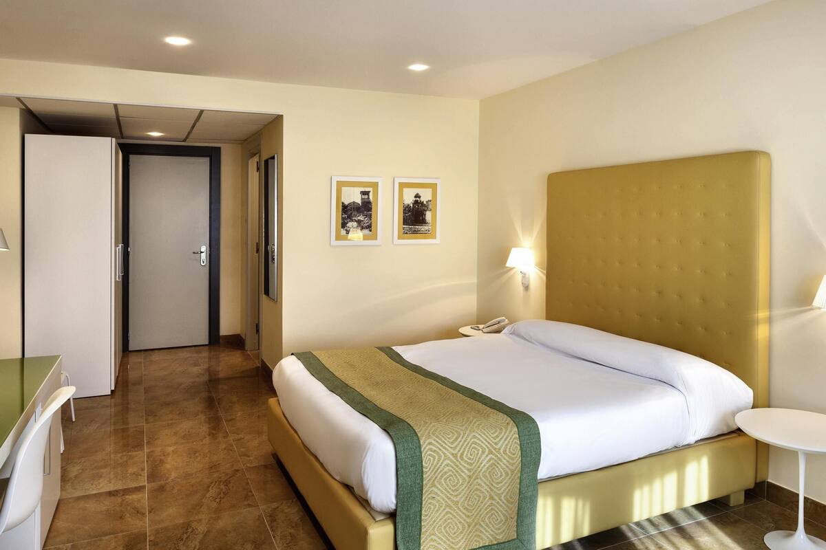 A hotel room with a bed, a desk, a chair, a bedside table, and two framed pictures on the wall.