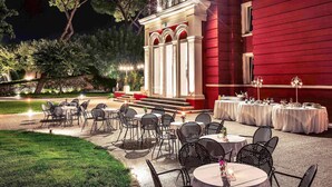 An outdoor dining area with tables and chairs, a red building with a balcony, and trees in the background. - Mercure Villa Romanazzi Carducci Bari (Bari)
