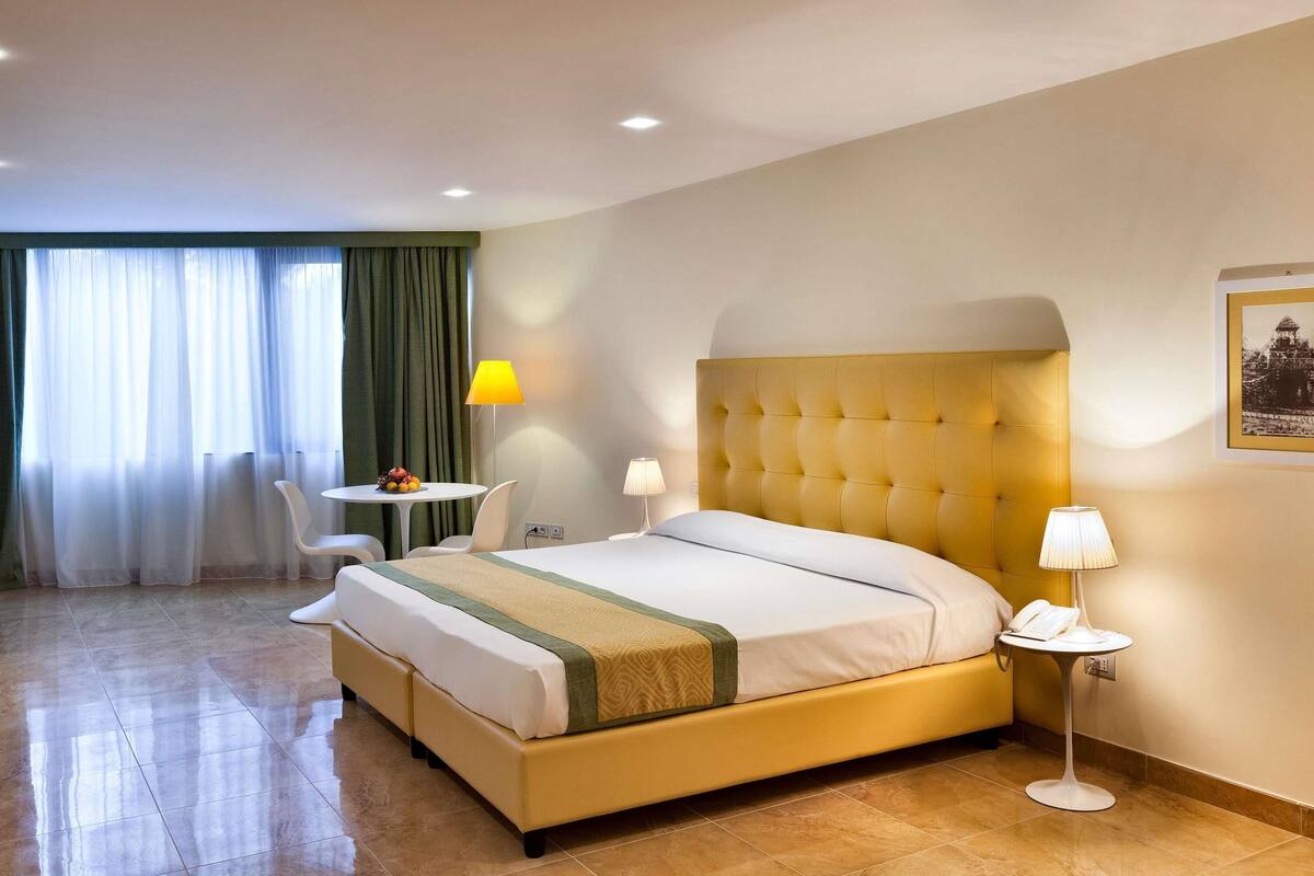 Una camera d'albergo con un letto, un tavolino, una sedia, una lampada e un quadro appeso al muro.