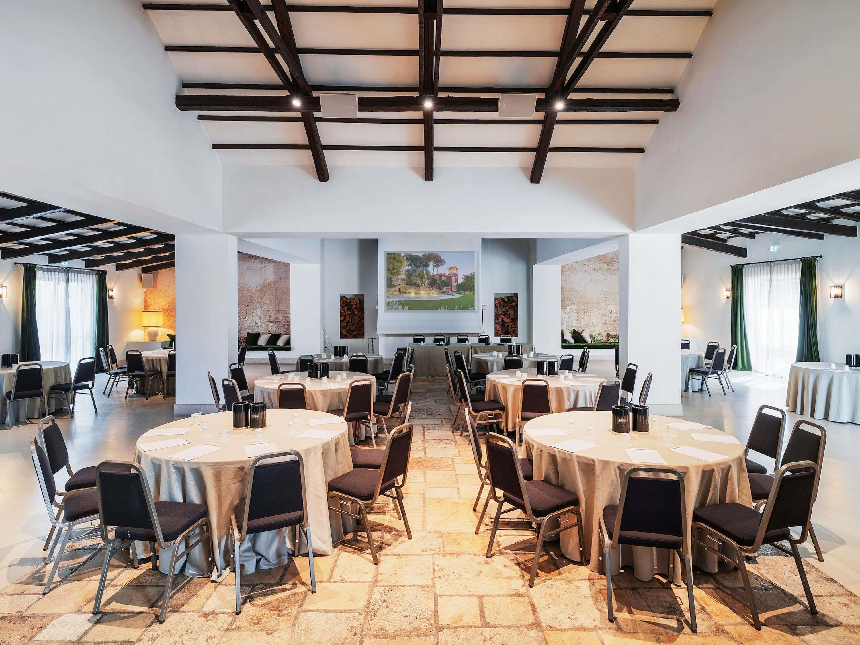 A spacious conference room with multiple round tables and chairs, a high ceiling with exposed beams, and artwork on the walls.
