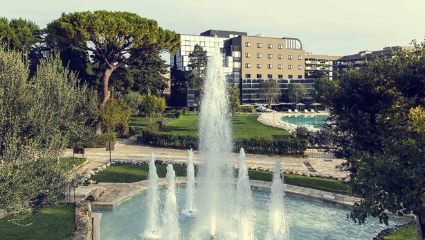 Une fontaine dans un parc, avec un bassin et un bâtiment en arrière-plan.