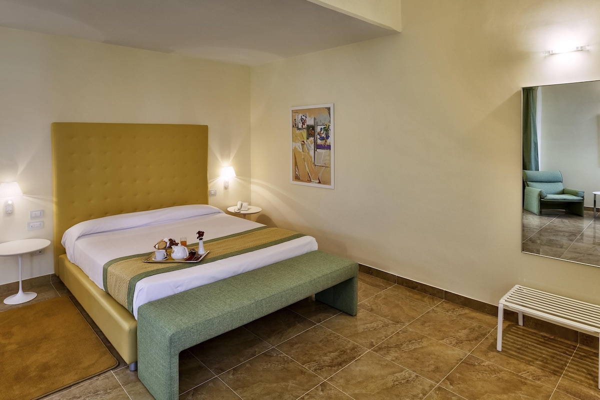 A hotel room with a bed, bedside table, lamp, bench, and a mirror.