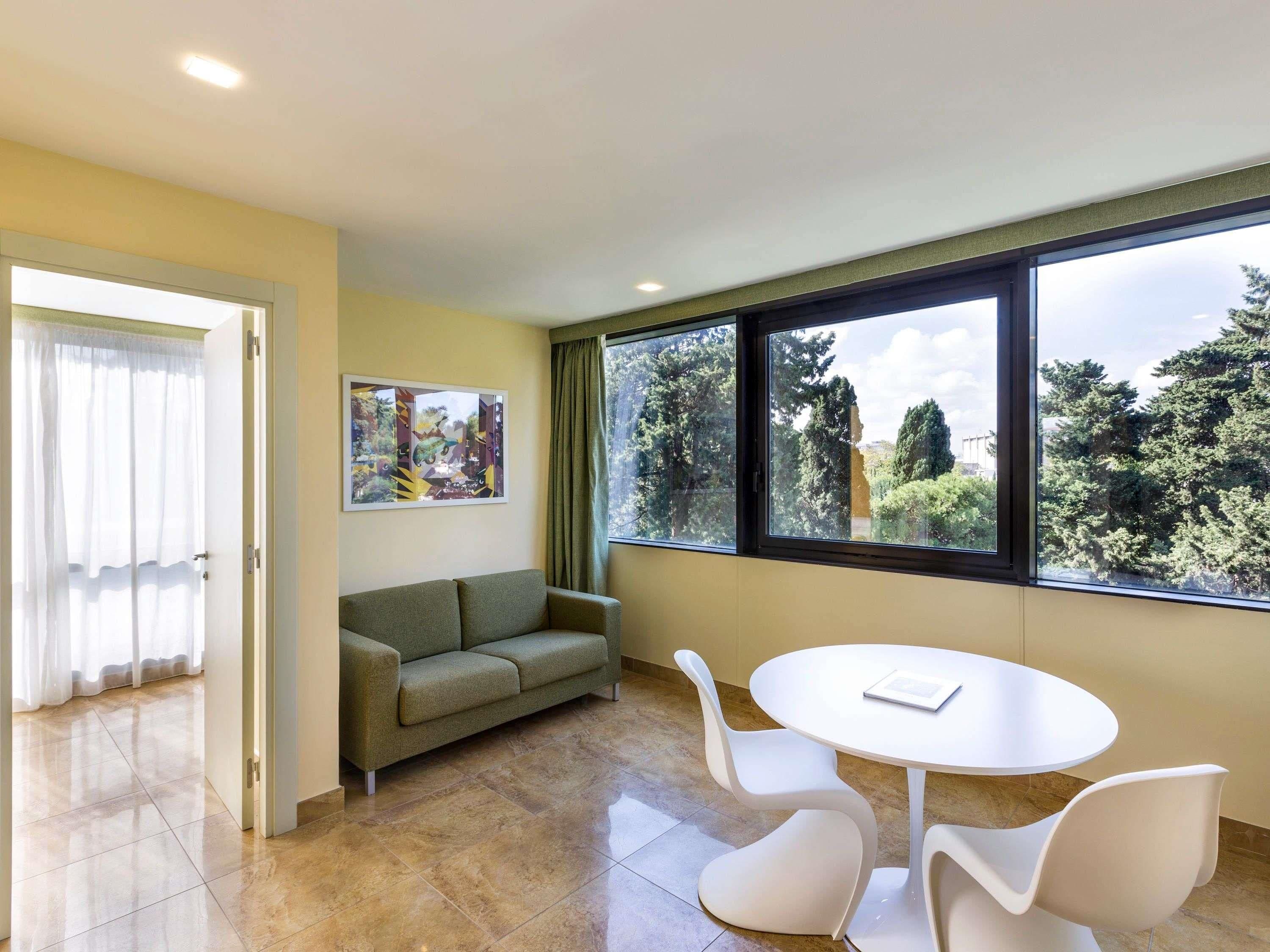 A modern room with a sofa, a round table, and chairs, a large window with a view of trees, and a painting on the wall.