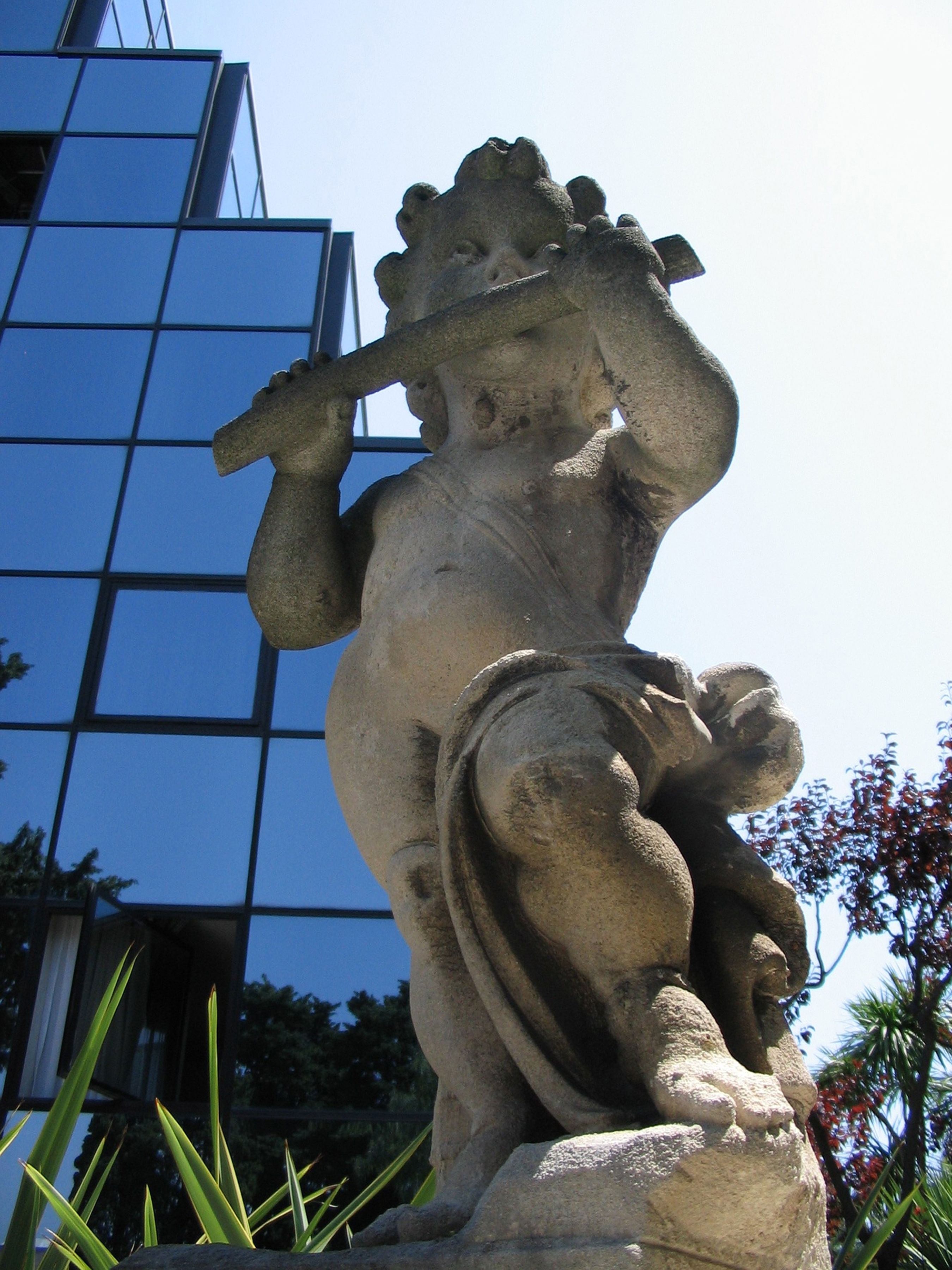 A stone statue of a figure holding a lyre, set against a modern building with glass windows.