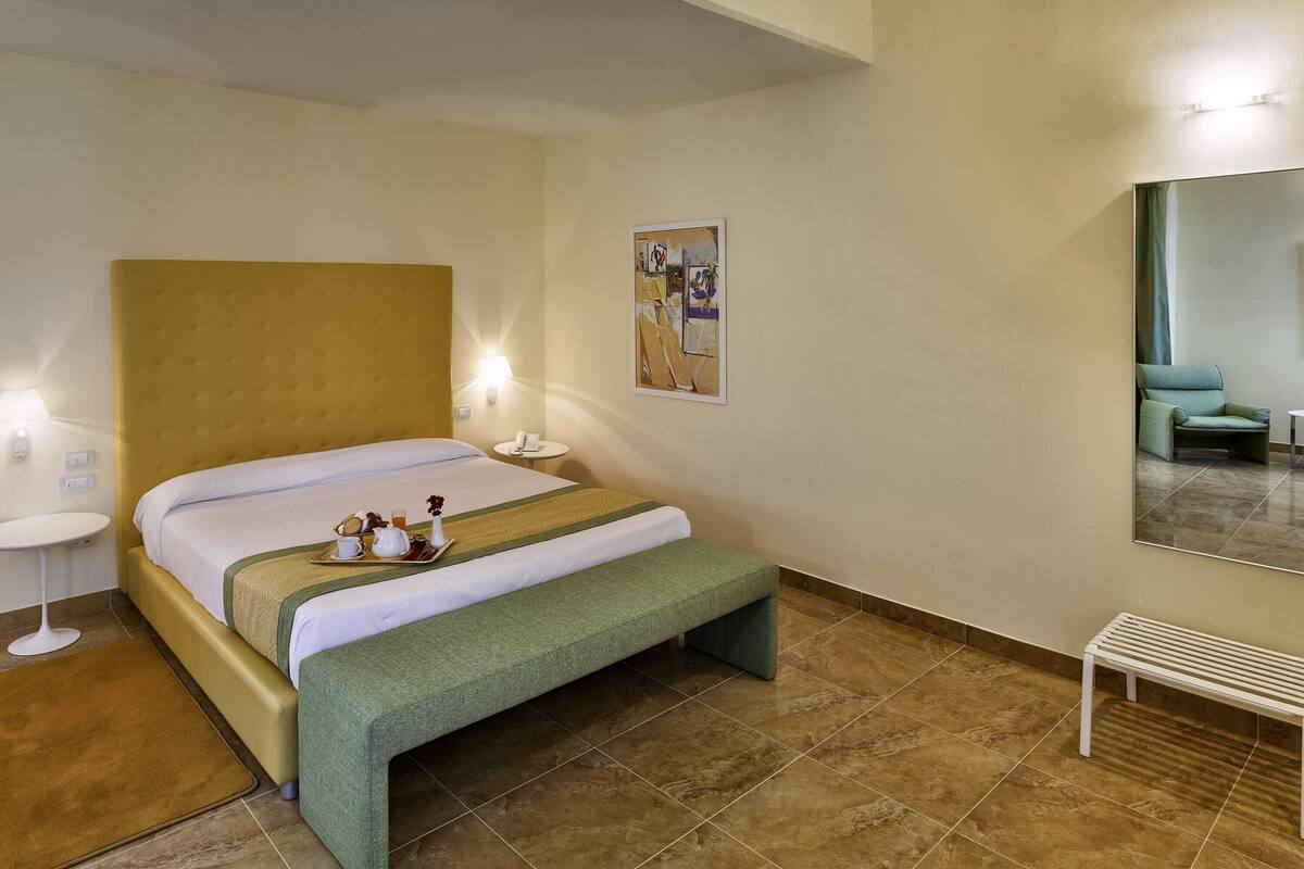 Una camera d'albergo con un letto, un comodino, una lampada, una panca e uno specchio.