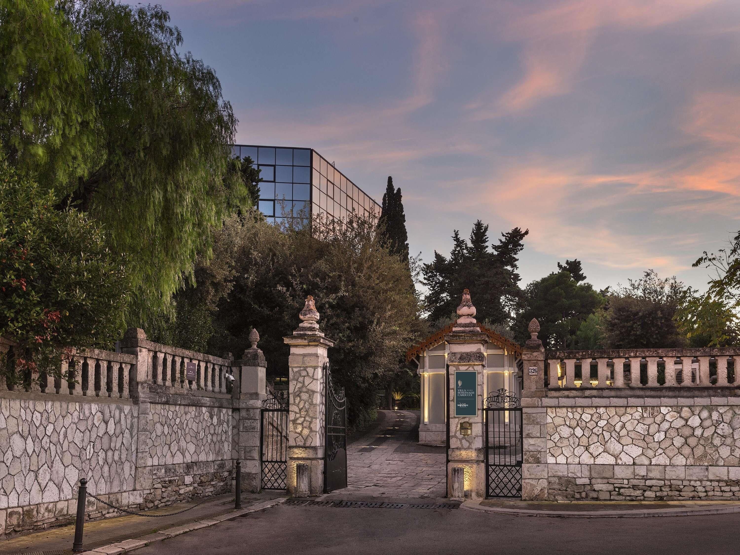A stone gate with a small shrine, a paved pathway, and a modern building in the background.