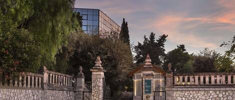 A stone gate with a small shrine, a paved pathway, and a modern building in the background.
