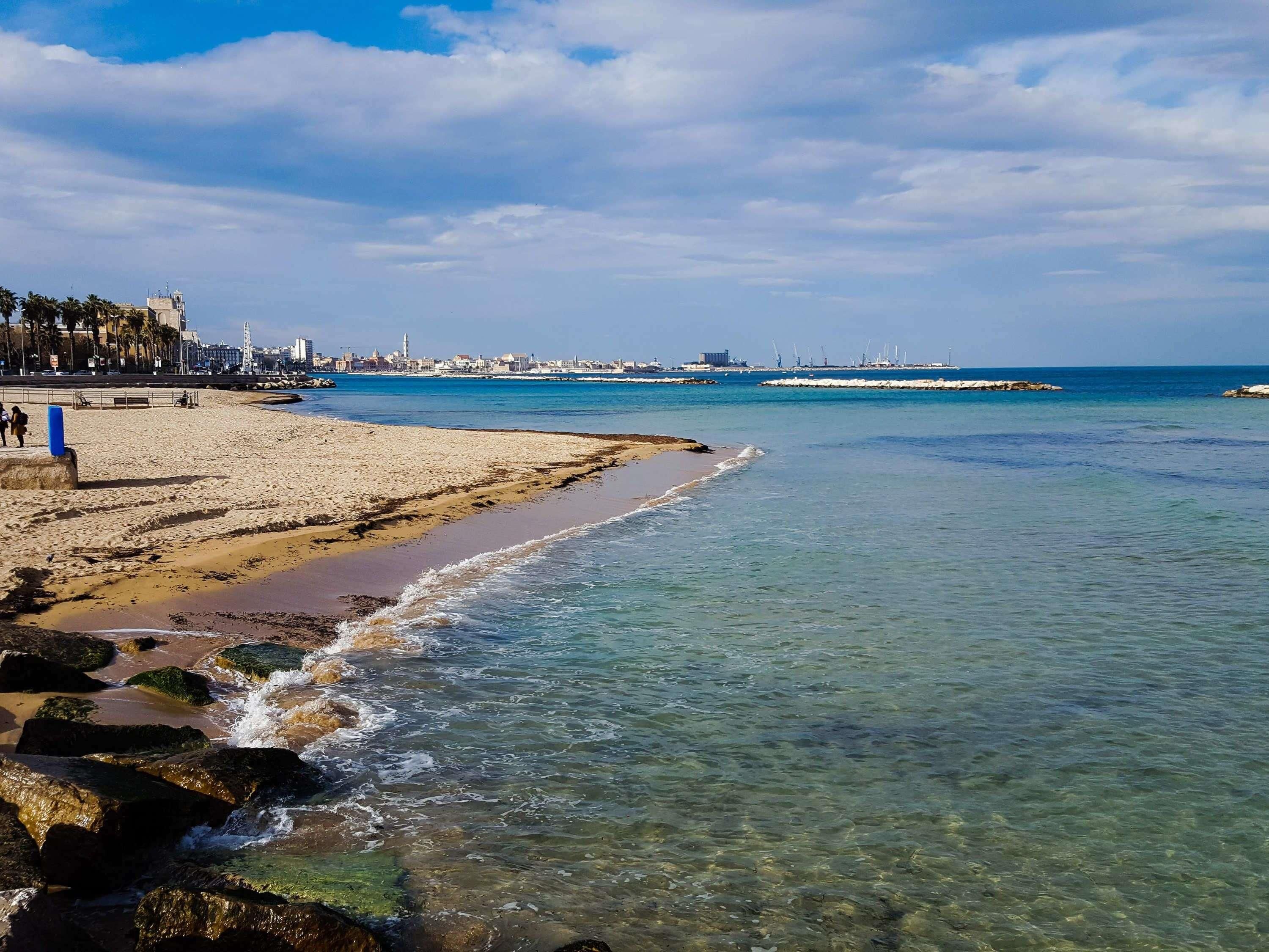A beach with clear water, a sandy shore, and a distant cityscape.