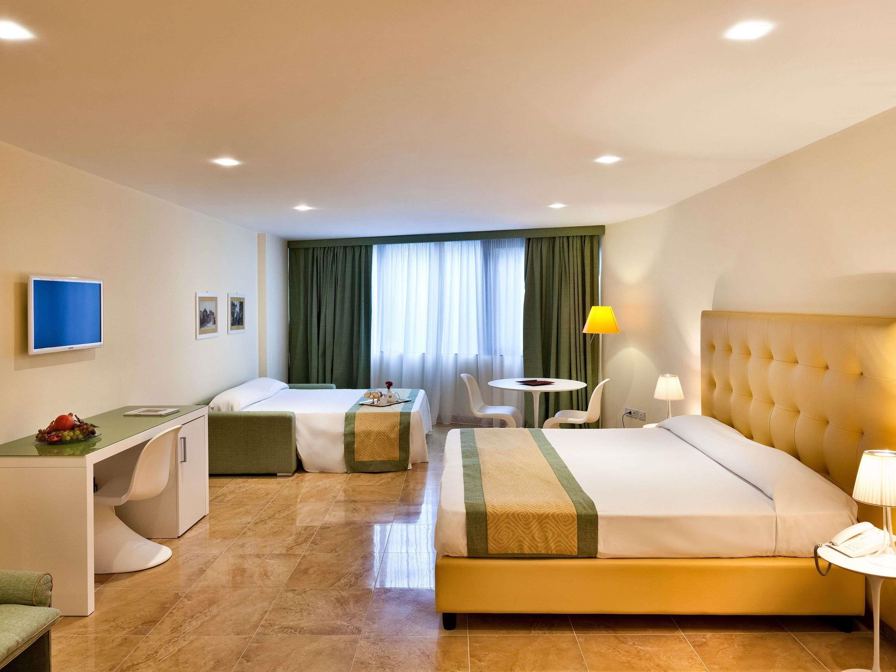 Ein Hotelzimmer mit Bett, Schreibtisch, Stuhl, kleinem Tisch, Fernseher und einem Fenster mit Vorhängen.