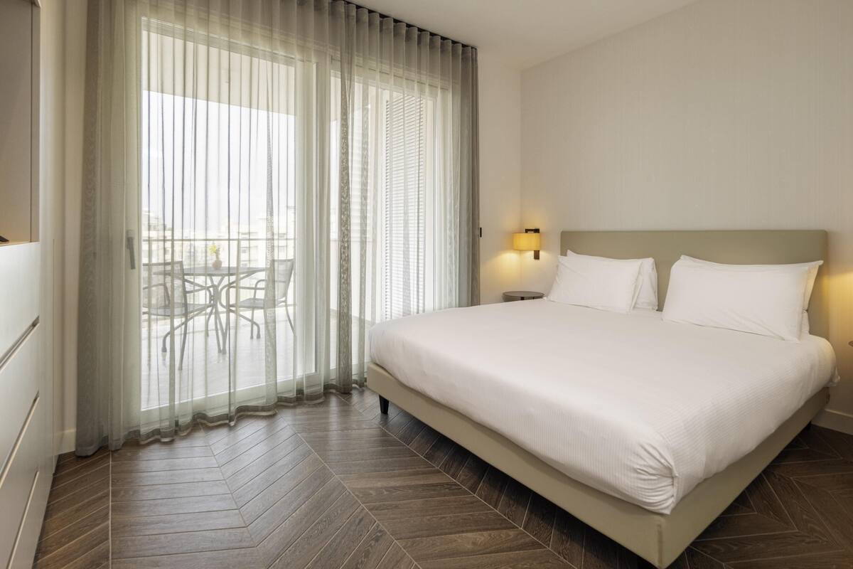 A hotel room with a large bed, a small table, and a view through sheer curtains.