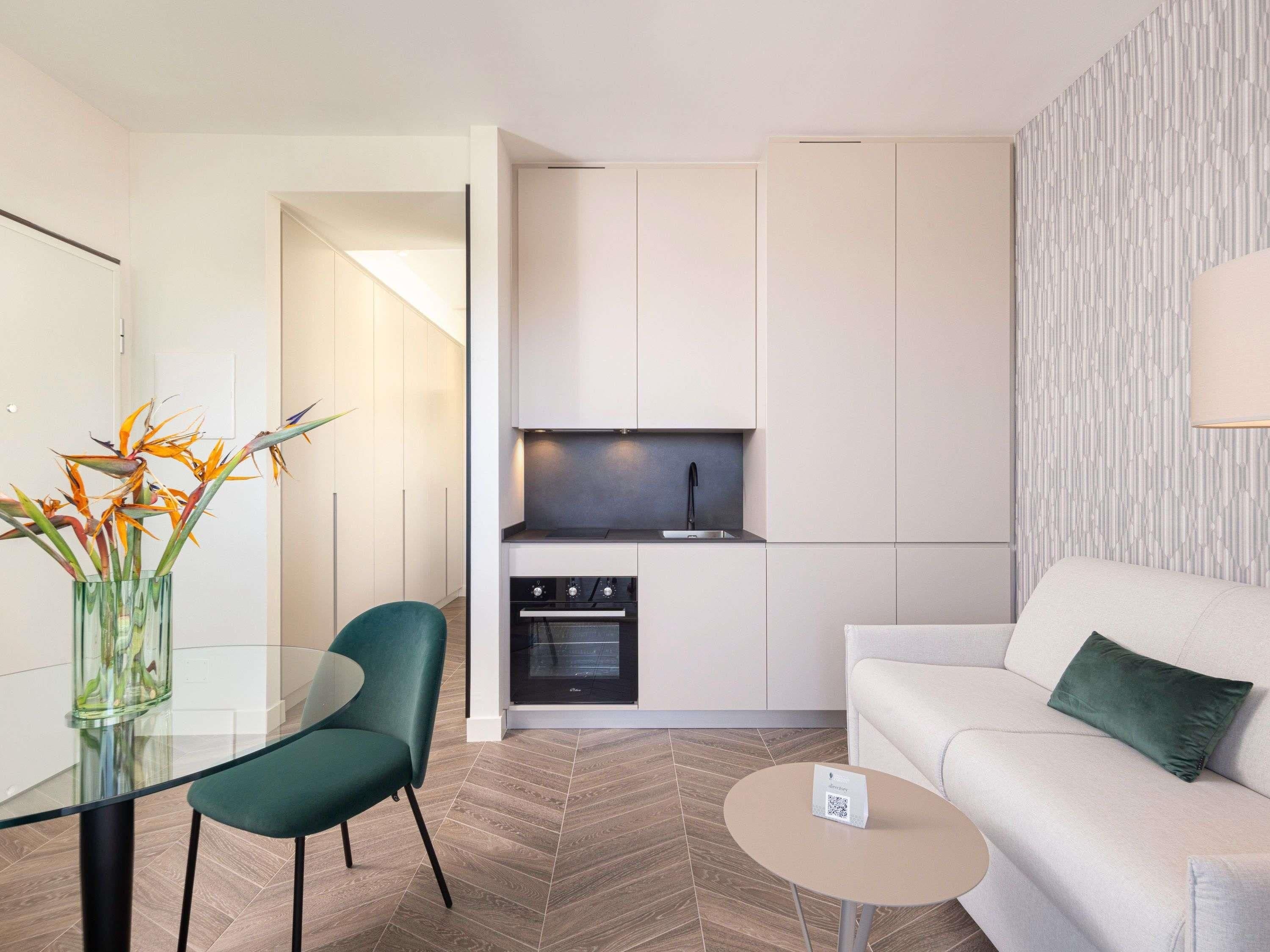 Un espace de vie compact comprenant un coin cuisine, un coin repas avec une table en verre, un canapé et un mur décoré de motifs.