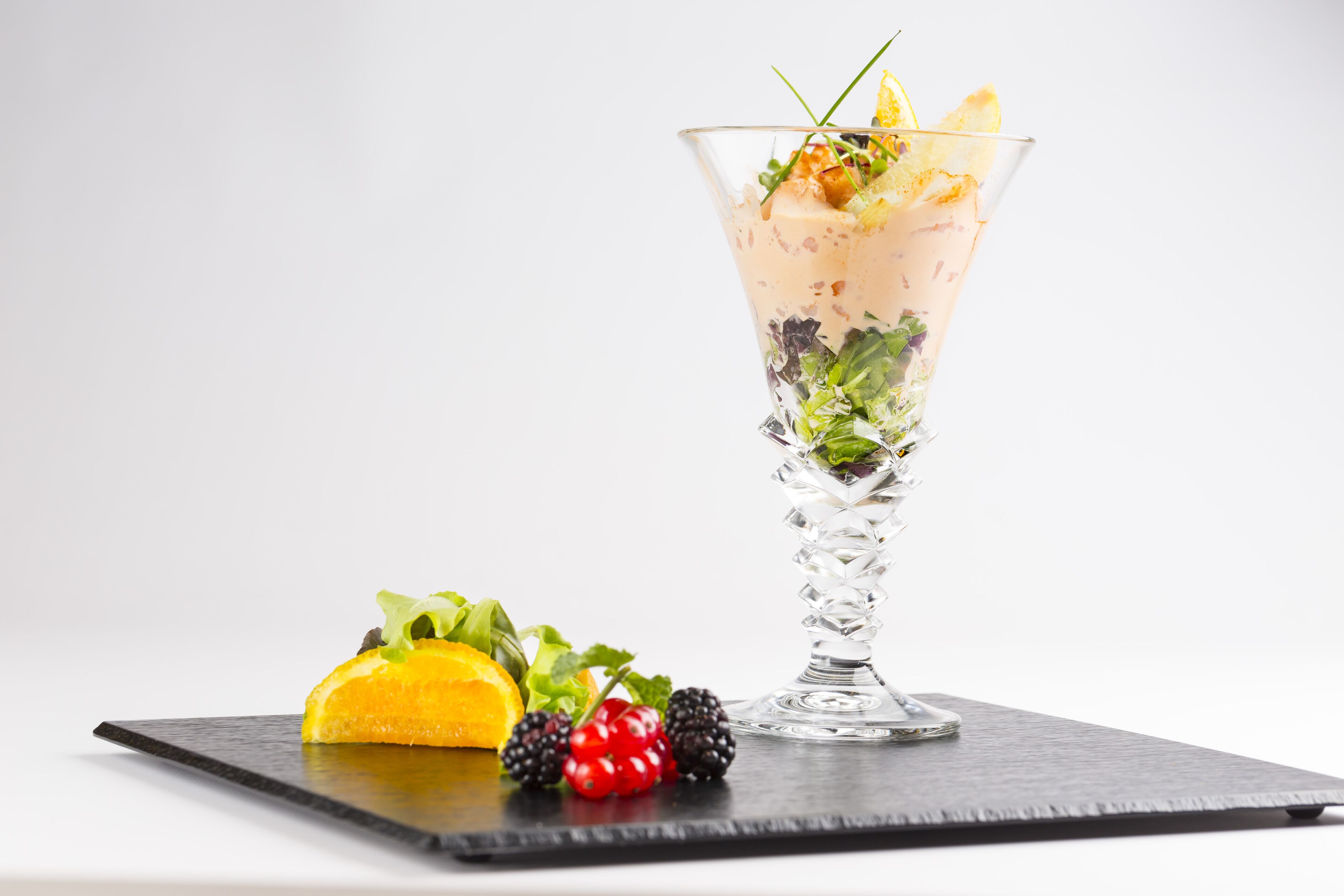 A cocktail glass with a layered drink, garnished with a lemon slice, herbs, and a cherry, accompanied by a slice of orange and blackberries on a slate serving tray.