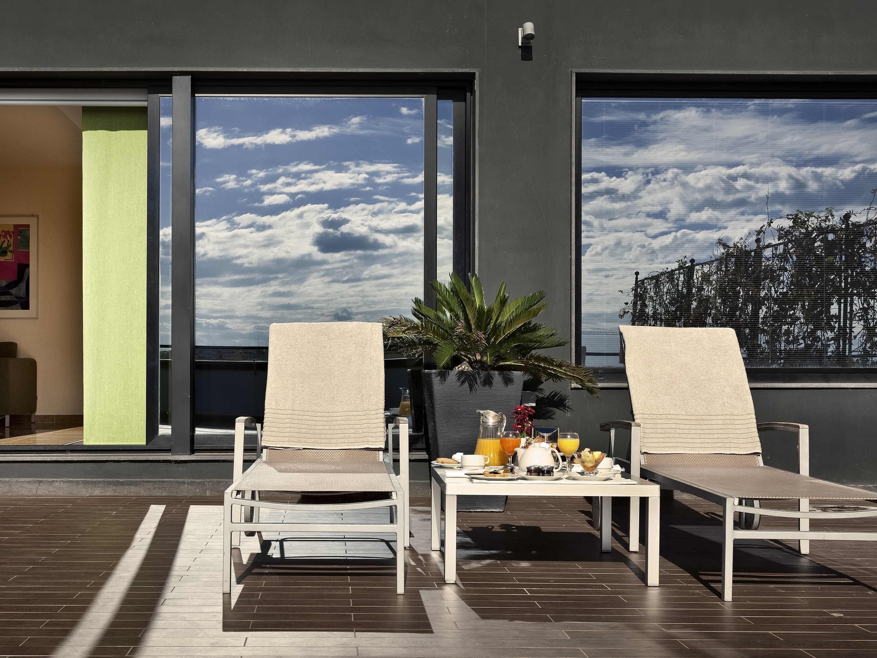 Two white lounge chairs with beige cushions on a tiled patio, a small table with breakfast items, and a large window with a view of the sky and clouds.