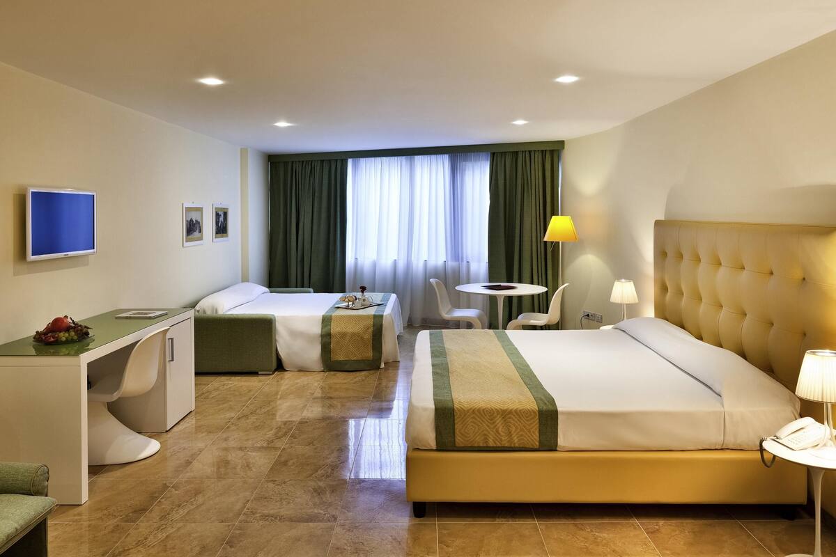 A hotel room with a bed, a desk, a chair, a small table, a TV, and a window with curtains.